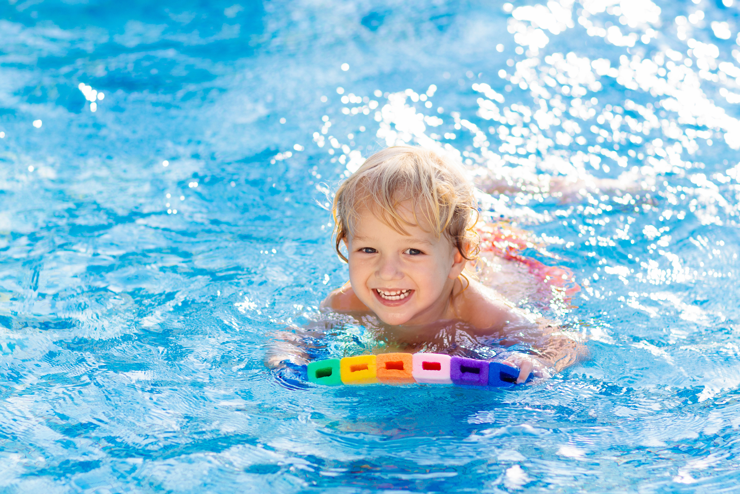 Child learning to swim in outdoor pool of tropical resort. Kids learn swimming. Exercise and training for young children. Little boy with colorful float board in sport club. Swimming baby or toddler.